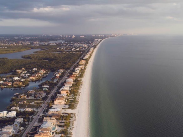 Stunning aerial view of coastal homes lining a serene beach with the ocean and clouds in the distance.